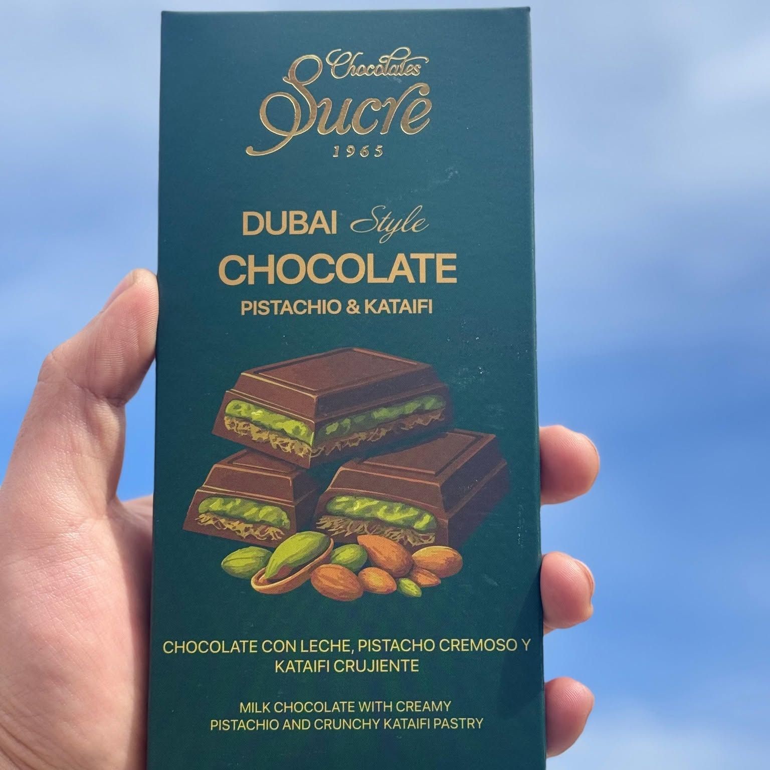 Una mano sostiene la caja verde de la tableta Estilo Dubái de Chocolates Sucre contra el cielo